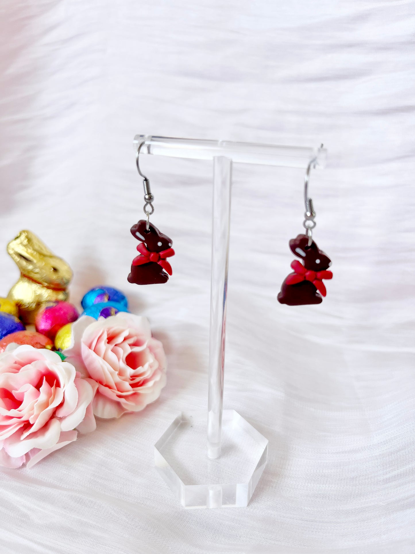 Silverstar.nzs Chocolate Bunny Polymer Clay Earrings, with red bunny shapes and small bows, hang on a clear stand. In the background, gold and red chocolate bunnies, colorful eggs, and pink roses are set against a white textured surface for a whimsical display of these funky earrings.