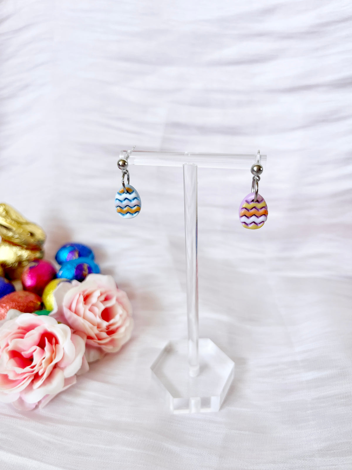 The Decorated Eggs Polymer Clay Earrings by silverstar.nz, featuring vibrant egg-shaped pendants with zigzag patterns, hang on a clear stand. Nearby are pink roses, small colorful eggs, and a gold rabbit decoration on soft white fabric.
