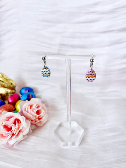 The Decorated Eggs Polymer Clay Earrings by silverstar.nz, featuring vibrant egg-shaped pendants with zigzag patterns, hang on a clear stand. Nearby are pink roses, small colorful eggs, and a gold rabbit decoration on soft white fabric.