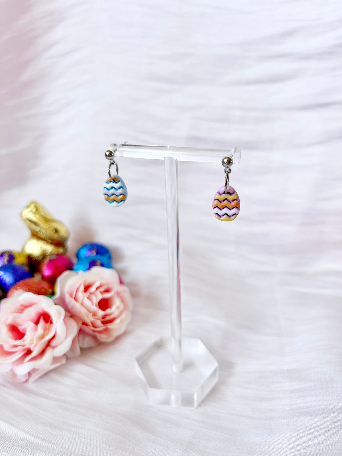 Decorated Eggs Polymer Clay Earrings by silverstar.nz, featuring colorful zigzag patterns and stainless steel hooks, hang on a transparent stand. Set against a white textured background, theyre accompanied by decorative roses and chocolate eggs, highlighting their unique design.