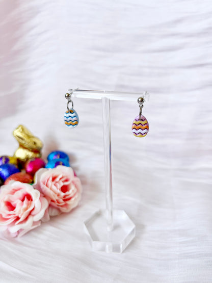 Decorated Eggs Polymer Clay Earrings by silverstar.nz, featuring colorful zigzag patterns and stainless steel hooks, hang on a transparent stand. Set against a white textured background, theyre accompanied by decorative roses and chocolate eggs, highlighting their unique design.
