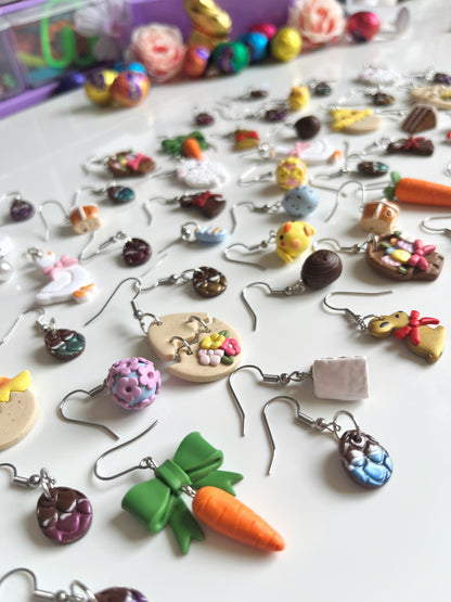 A colorful set of silverstar.nzs handmade polymer clay earrings is displayed on a white surface. The unique designs include Carrots with a Bow, cookies, and chicks, featuring a playful, crafty theme. Blurred colorful objects are visible in the background.