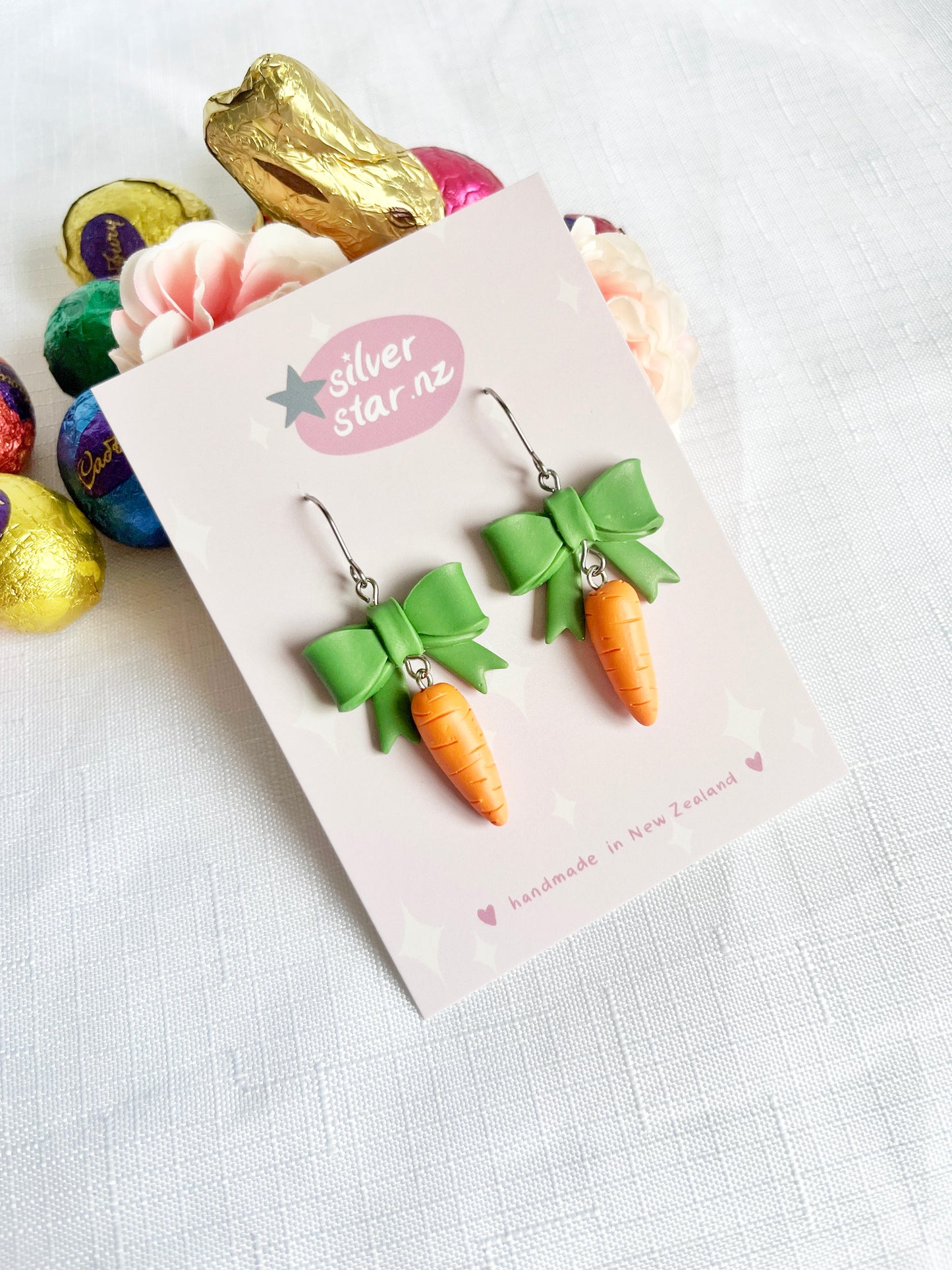 The Carrots with a Bow Polymer Clay Earrings from silverstar.nz feature on a white display card amidst colorful chocolates and roses, offering a charming handmade design of small carrots with green bows.