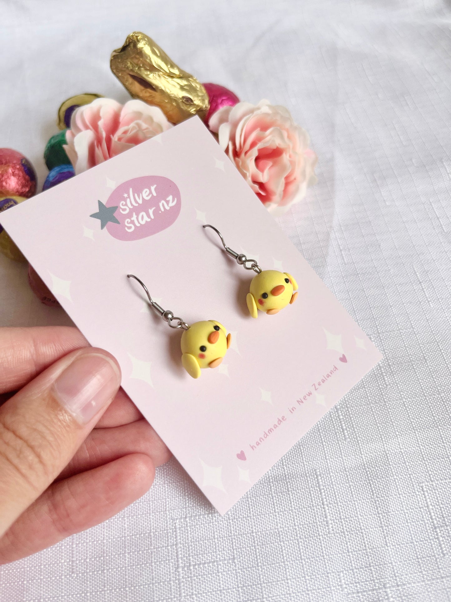 Displayed on a silverstar.nz branded card, the Mini Baby Chick Polymer Clay Earrings showcase small, round yellow charms. In the background lie vibrant Easter eggs, pink roses, and a gold-hued rabbit on a textured surface. Enjoy the charm of this unique design.