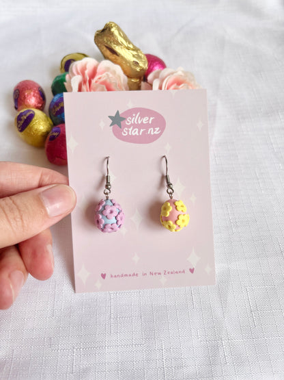 A hand displays Floral Egg Polymer Clay Earrings by silverstar.nz, shaped like pink and yellow eggs with floral designs. The card reads handmade in New Zealand, featuring durable stainless steel hooks. The background is decorated with colorful eggs and pink flowers on a white cloth.