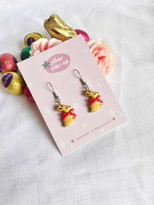Gold Bunny Polymer Clay Earrings by silverstar.nz are handmade and shaped like gold chocolate bunnies with red ribbons, showcased on a branded card, surrounded by pastel roses and colorful chocolate eggs on a white textured background.