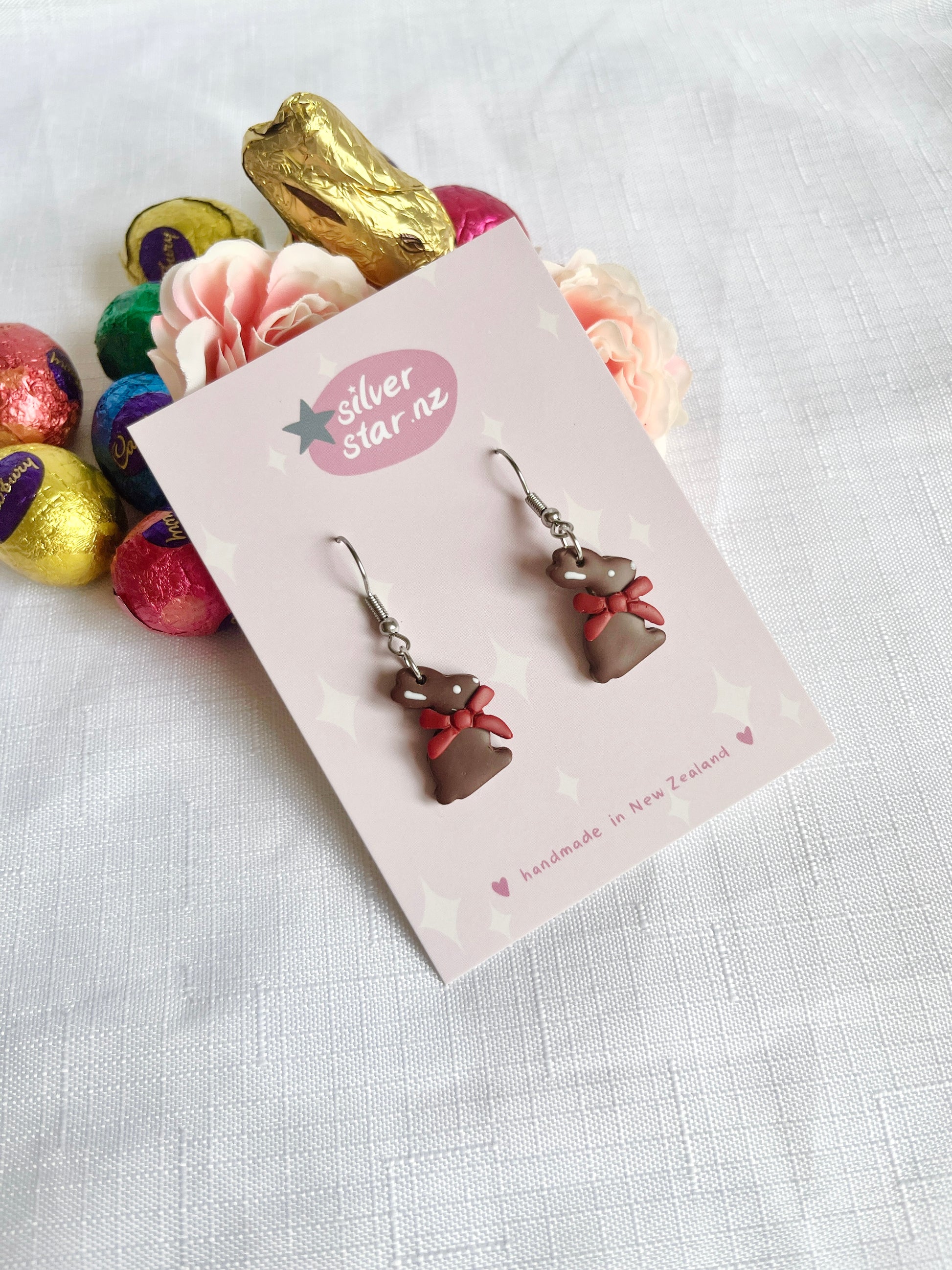 A pair of Chocolate Bunny Polymer Clay Earrings by silverstar.nz, featuring a funky bear-shaped design with red bows on a card, set against white fabric with colorful wrapped chocolate eggs and a large chocolate bunny in the background.