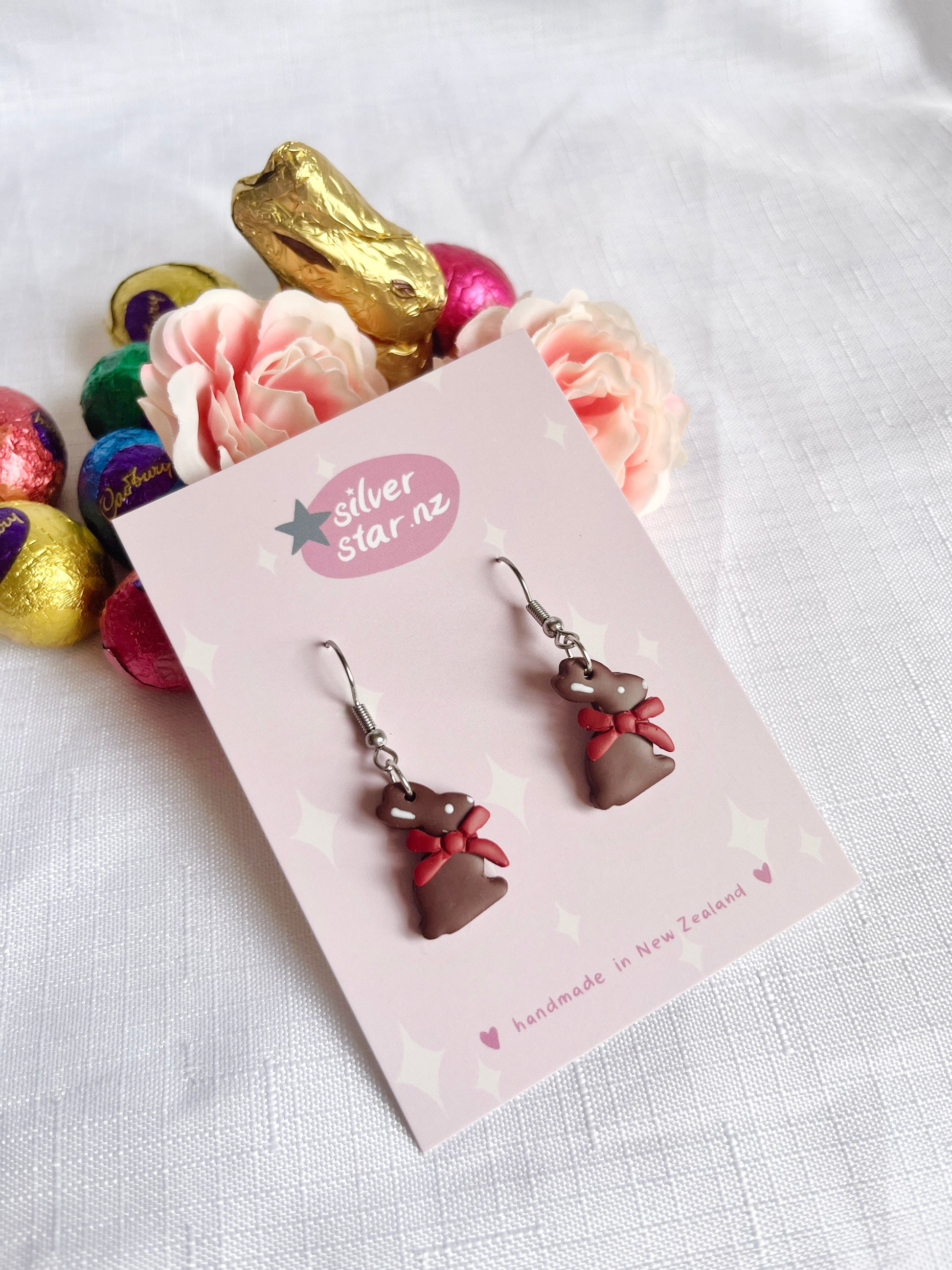 Cute handmade Chocolate Bunny Polymer Clay Earrings by silverstar.nz, featuring brown bunnies with red bows, are displayed on a card surrounded by roses and colorful chocolate eggs, adding a funky design flair to your accessory collection.