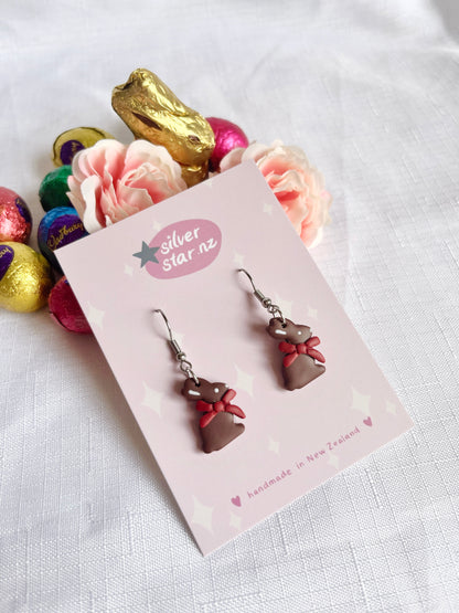 Cute handmade Chocolate Bunny Polymer Clay Earrings by silverstar.nz, featuring brown bunnies with red bows, are displayed on a card surrounded by roses and colorful chocolate eggs, adding a funky design flair to your accessory collection.