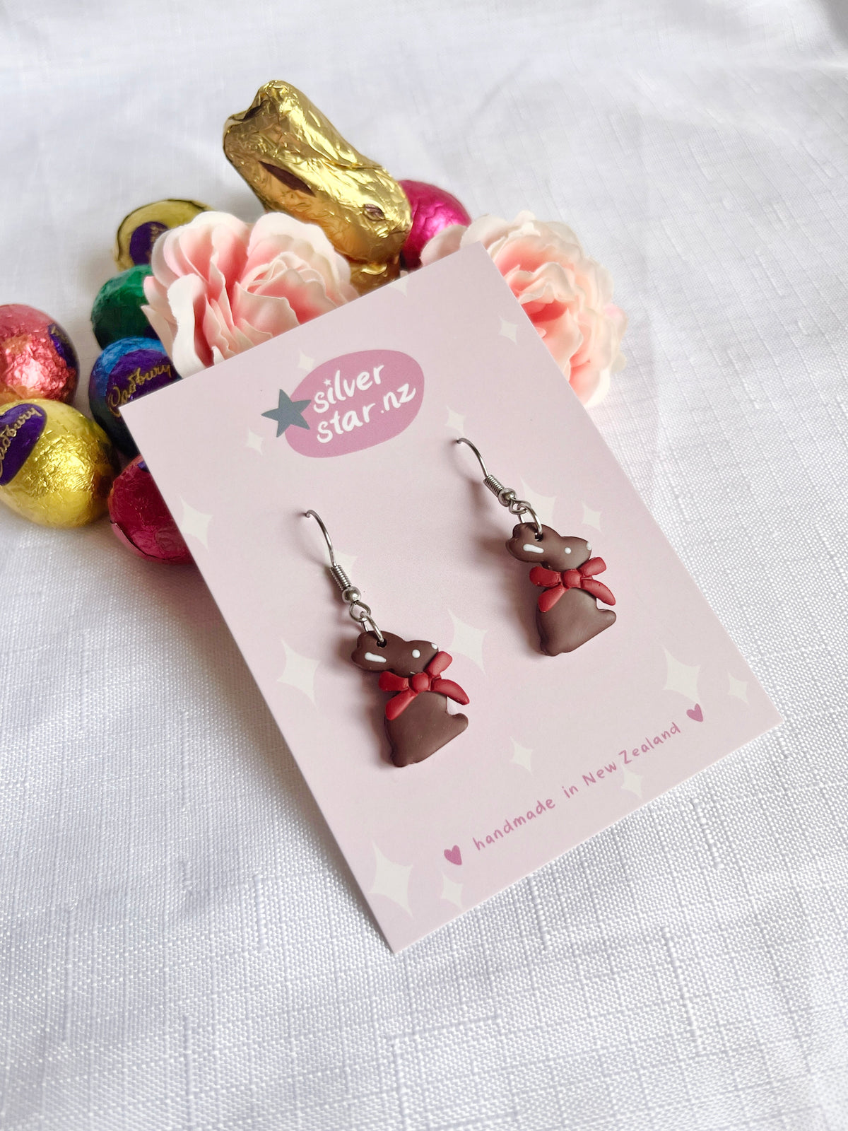 The handmade Chocolate Bunny Polymer Clay Earrings by silverstar.nz, featuring red bows, are displayed on a branded card against white fabric. A pink flower, gold chocolate bunny, and colorful foil-wrapped eggs add funky design flair in the backdrop.