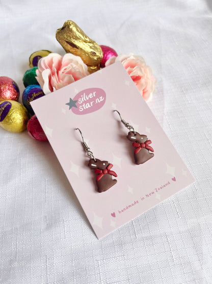 The handmade Chocolate Bunny Polymer Clay Earrings by silverstar.nz, featuring red bows, are displayed on a branded card against white fabric. A pink flower, gold chocolate bunny, and colorful foil-wrapped eggs add funky design flair in the backdrop.