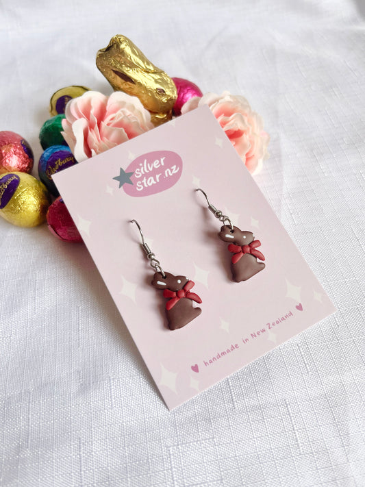 The handmade Chocolate Bunny Polymer Clay Earrings by silverstar.nz, featuring red bows, are displayed on a branded card against white fabric. A pink flower, gold chocolate bunny, and colorful foil-wrapped eggs add funky design flair in the backdrop.