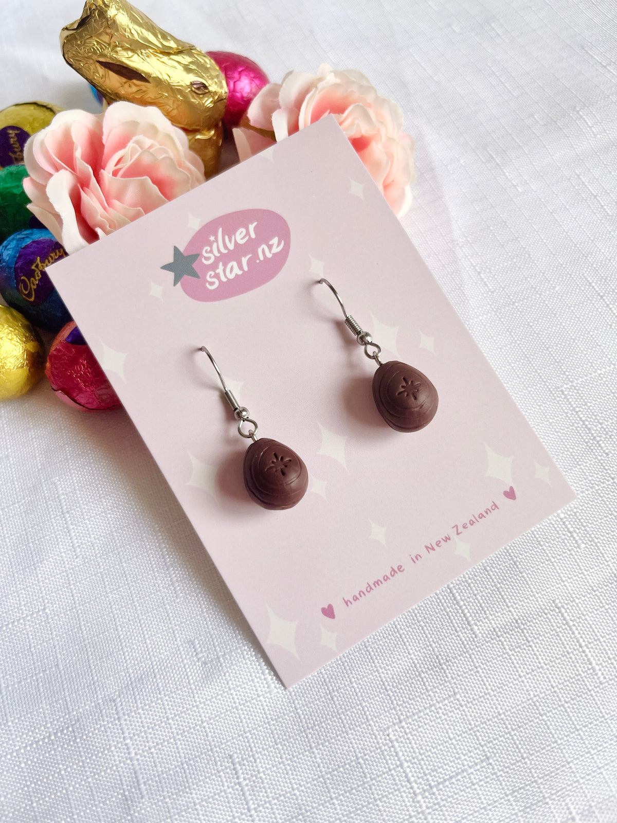 A pair of Mini Creme Egg Polymer Clay Earrings from silverstar.nz is displayed on a pink card. The setting features assorted colorful foil-wrapped chocolates and artificial pink roses, enhancing the charm of this delightful design against the white textured surface.