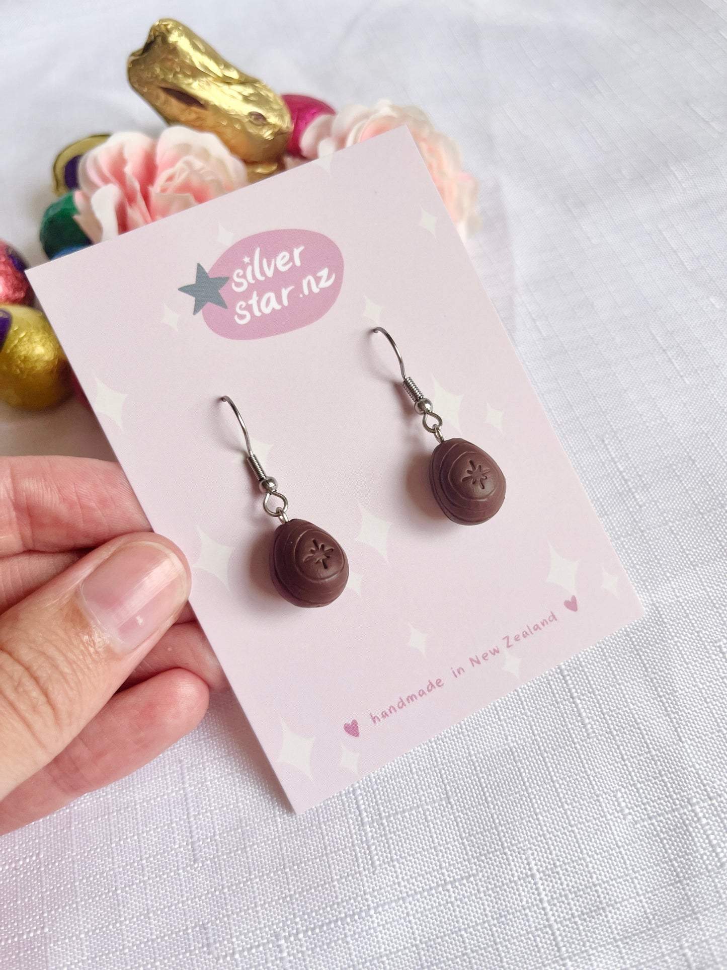 A hand holds an earring card labeled silverstar.nz, showcasing unique, handmade Mini Creme Egg Polymer Clay Earrings. The card, adorned with pastel stars, reads handmade in New Zealand. Behind it are polymer clay heart-shaped chocolates and pink flowers on a white cloth.