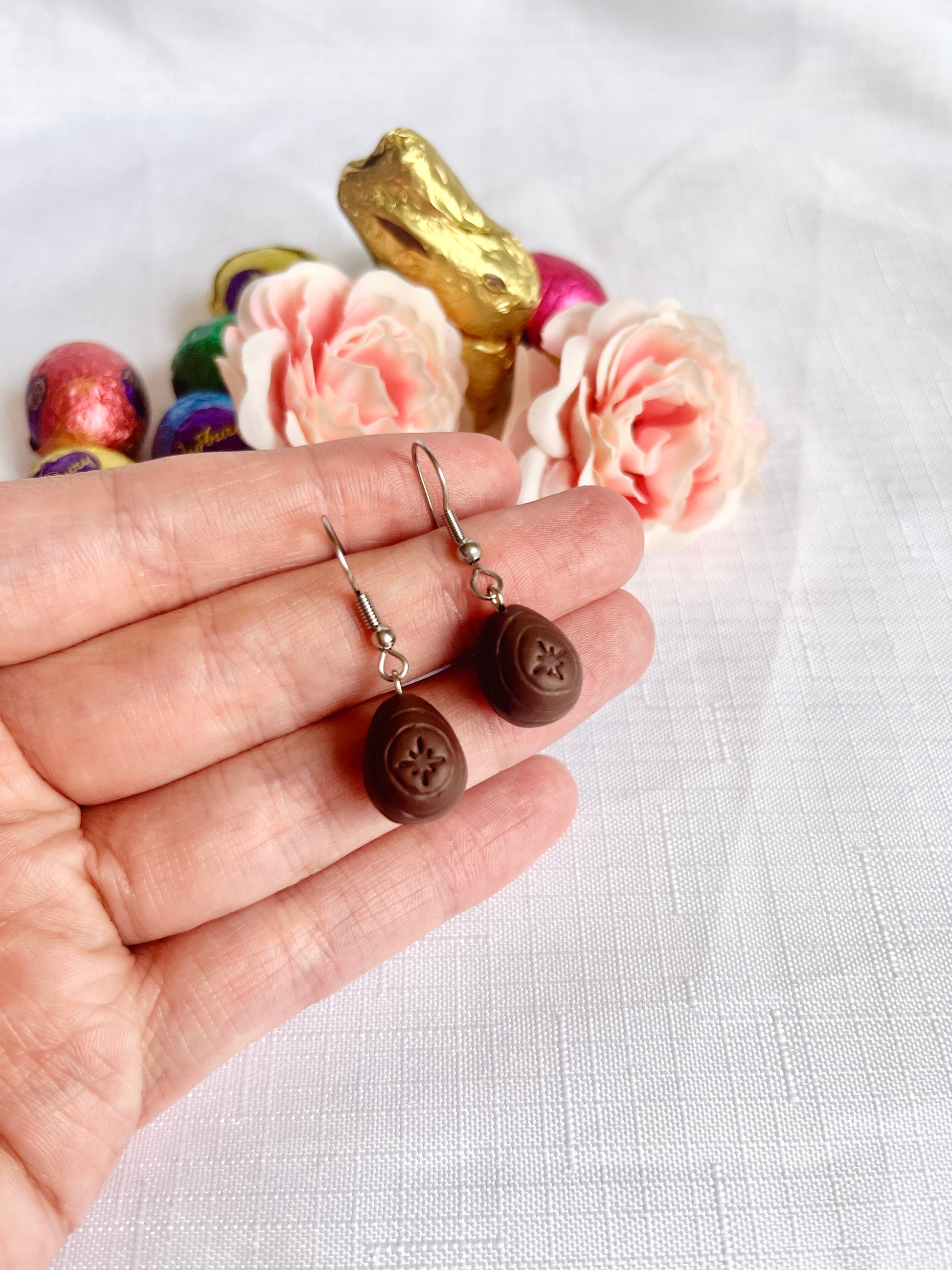 A hand holds a whimsical pair of silverstar.nz Mini Creme Egg Polymer Clay Earrings. The background features pink roses, colorful foil-wrapped chocolate eggs, and a golden chocolate bunny on a white surface—a truly unique design for fans of playful elegance.