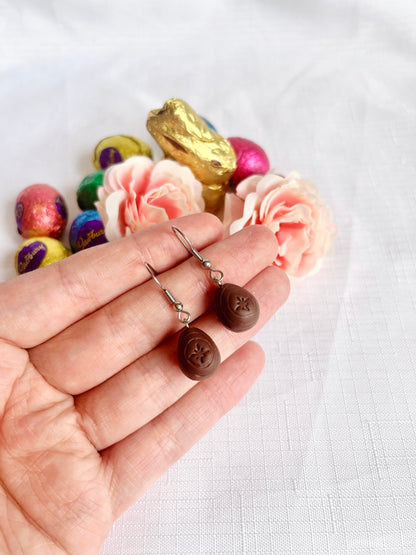 A hand holds a pair of Mini Creme Egg Polymer Clay Earrings by silverstar.nz, uniquely handmade with flower designs. In the background, assorted chocolate eggs and pink roses are artfully arranged on a white surface.
