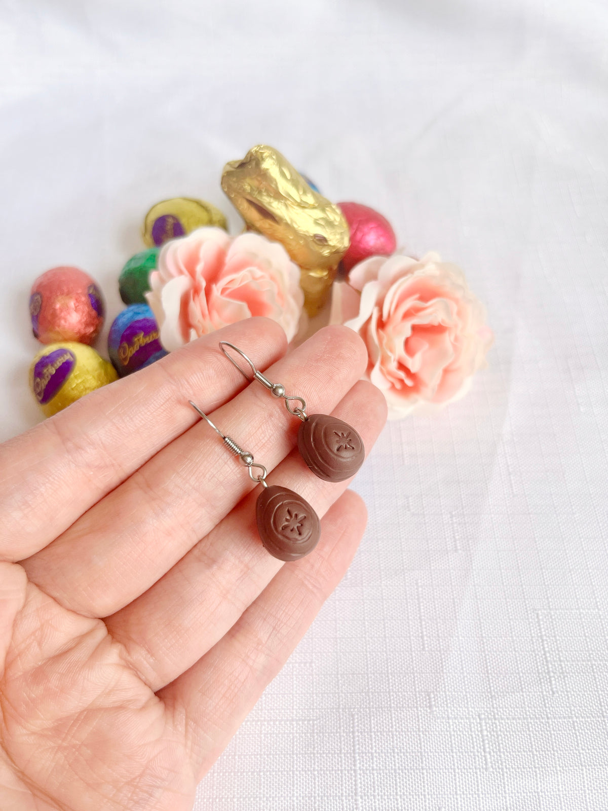 A hand holds a pair of silverstar.nzs Mini Creme Egg Polymer Clay Earrings, nestled among colorful chocolate eggs, a golden chocolate bunny, and soft pink roses on a white textured surface.