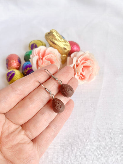 A hand holds a pair of silverstar.nzs Mini Creme Egg Polymer Clay Earrings, nestled among colorful chocolate eggs, a golden chocolate bunny, and soft pink roses on a white textured surface.