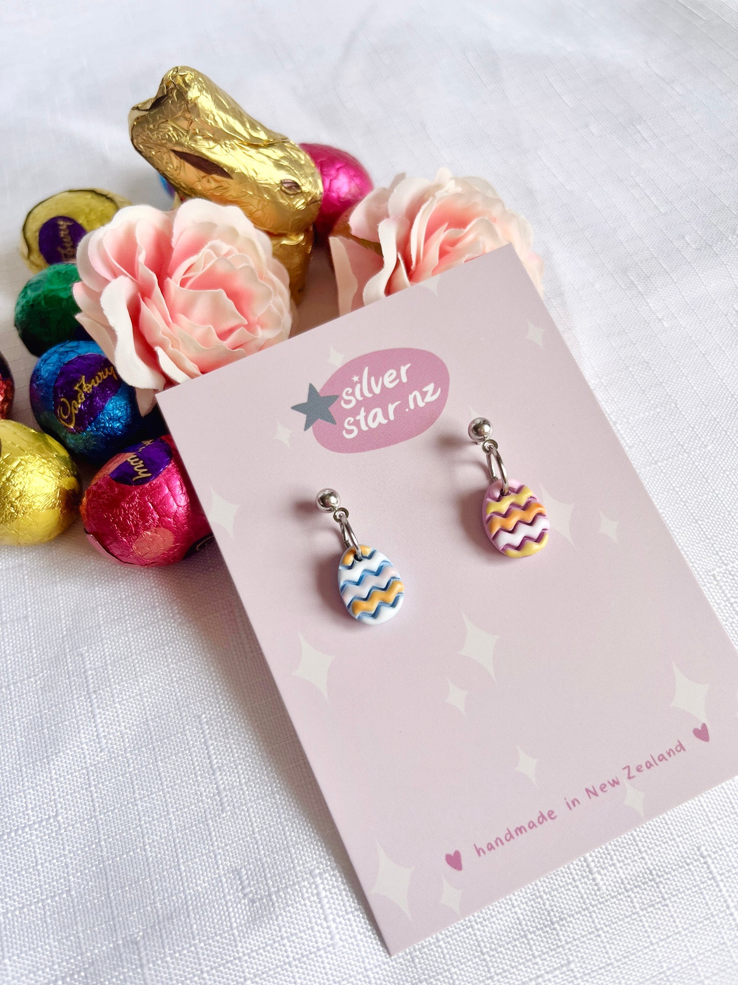 The Decorated Eggs Polymer Clay Earrings by silverstar.nz feature a unique funky design with zigzag patterns in pink, blue, and yellow on Easter egg shapes. Displayed on a silverstar.nz card, they include stainless steel hooks and are surrounded by roses and colorful foil-wrapped chocolates.