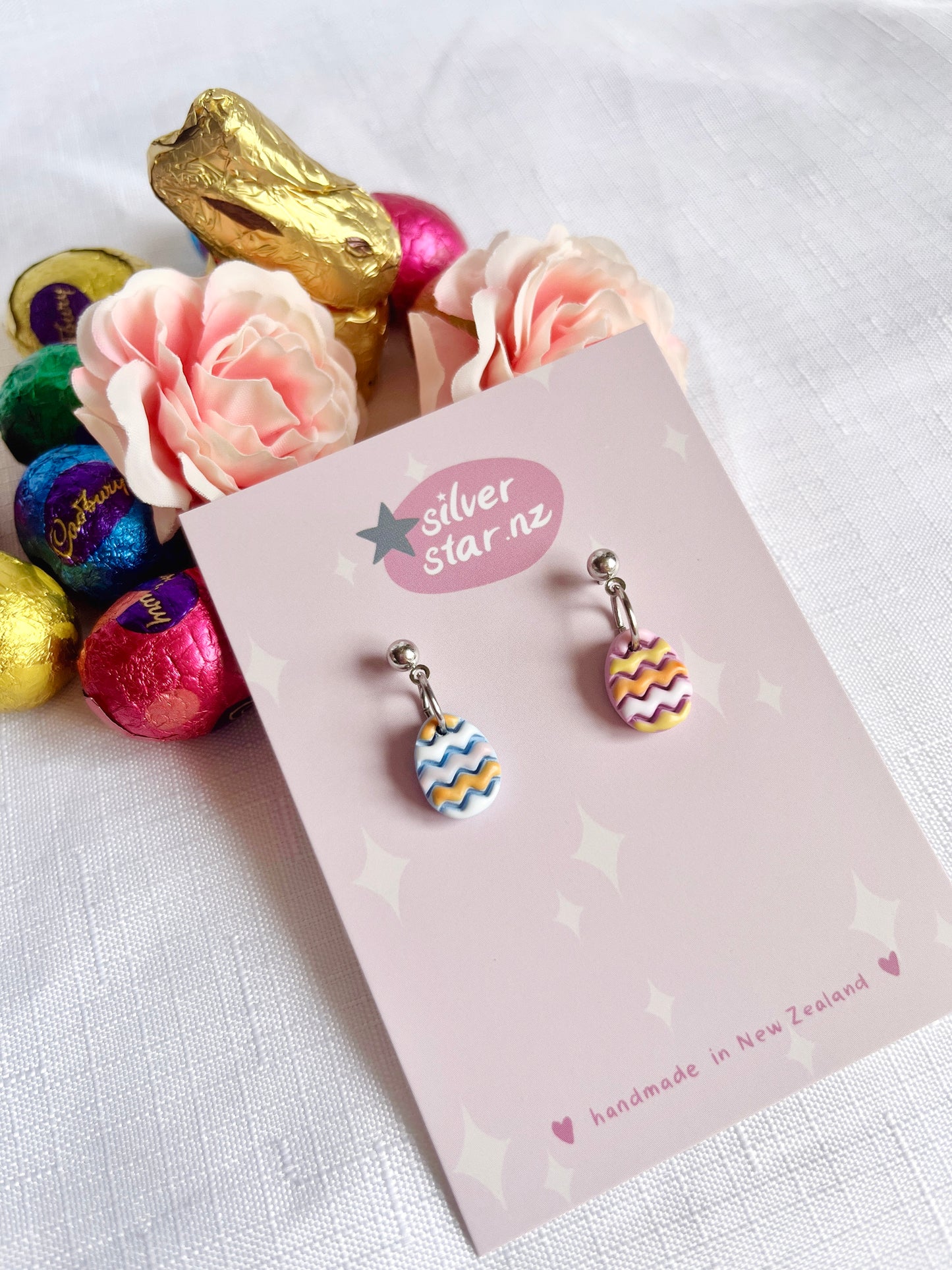 A display card for Decorated Eggs Polymer Clay Earrings by silverstar.nz features unique handmade earrings with two dangling Easter egg designs—one in blue and white, the other in pink and yellow. The earrings hang from stainless steel hooks. Nearby are pink roses, a gold bunny, and colorful chocolates.