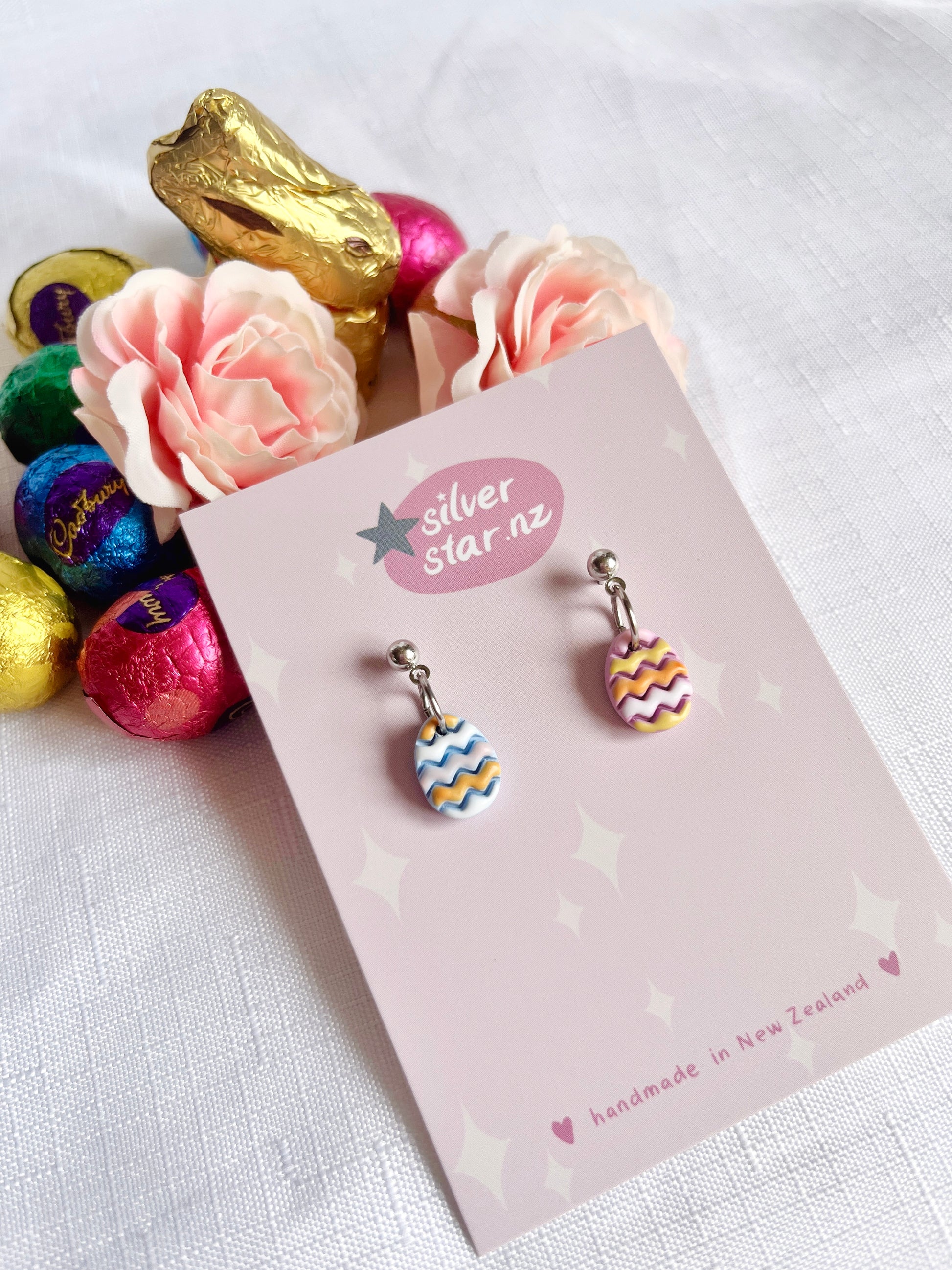 A display card for Decorated Eggs Polymer Clay Earrings by silverstar.nz features unique handmade earrings with two dangling Easter egg designs—one in blue and white, the other in pink and yellow. The earrings hang from stainless steel hooks. Nearby are pink roses, a gold bunny, and colorful chocolates.
