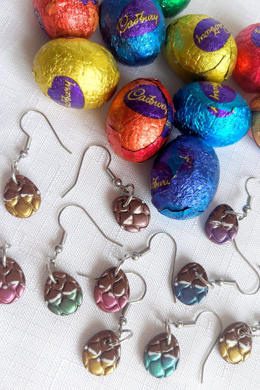 The Opened Easter Egg Polymer Clay Earrings by silverstar.nz, designed to resemble small, colorful chocolate eggs, are scattered on a white background. These handmade accessories mimic wrapped chocolate eggs in vibrant hues shown at the top of the image.