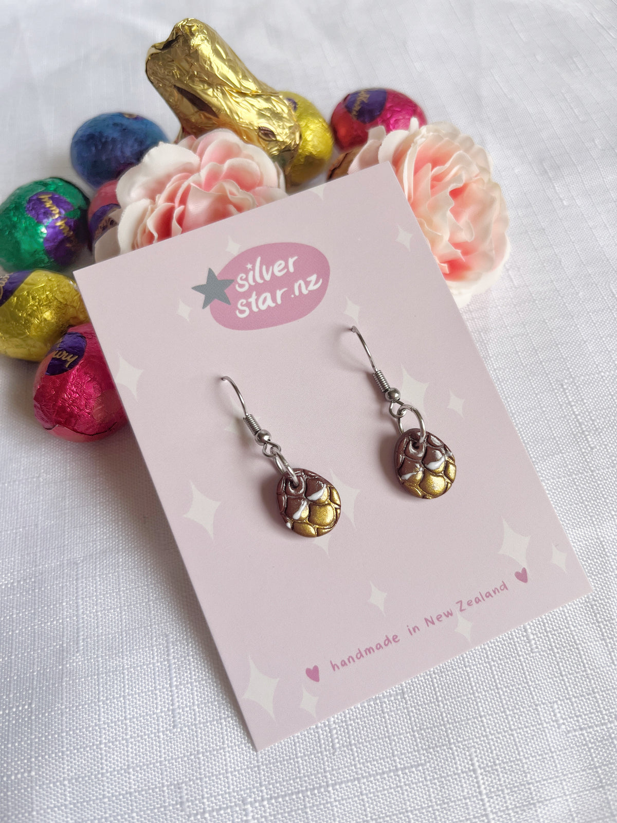 Displayed on a card labeled silverstar.nz, handmade in New Zealand, the Opened Easter Egg Polymer Clay Earrings feature a unique design. Surrounded by colorful chocolates and pink flowers on a white textured surface, these handmade earrings are truly exquisite.