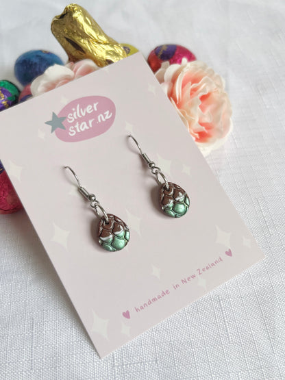 The Opened Easter Egg Polymer Clay earrings, teardrop-shaped with intricate, unique designs, are displayed on a card labeled silverstar.nz, handmade in New Zealand, against a backdrop of colorful wrapped chocolates and soft pink flowers.