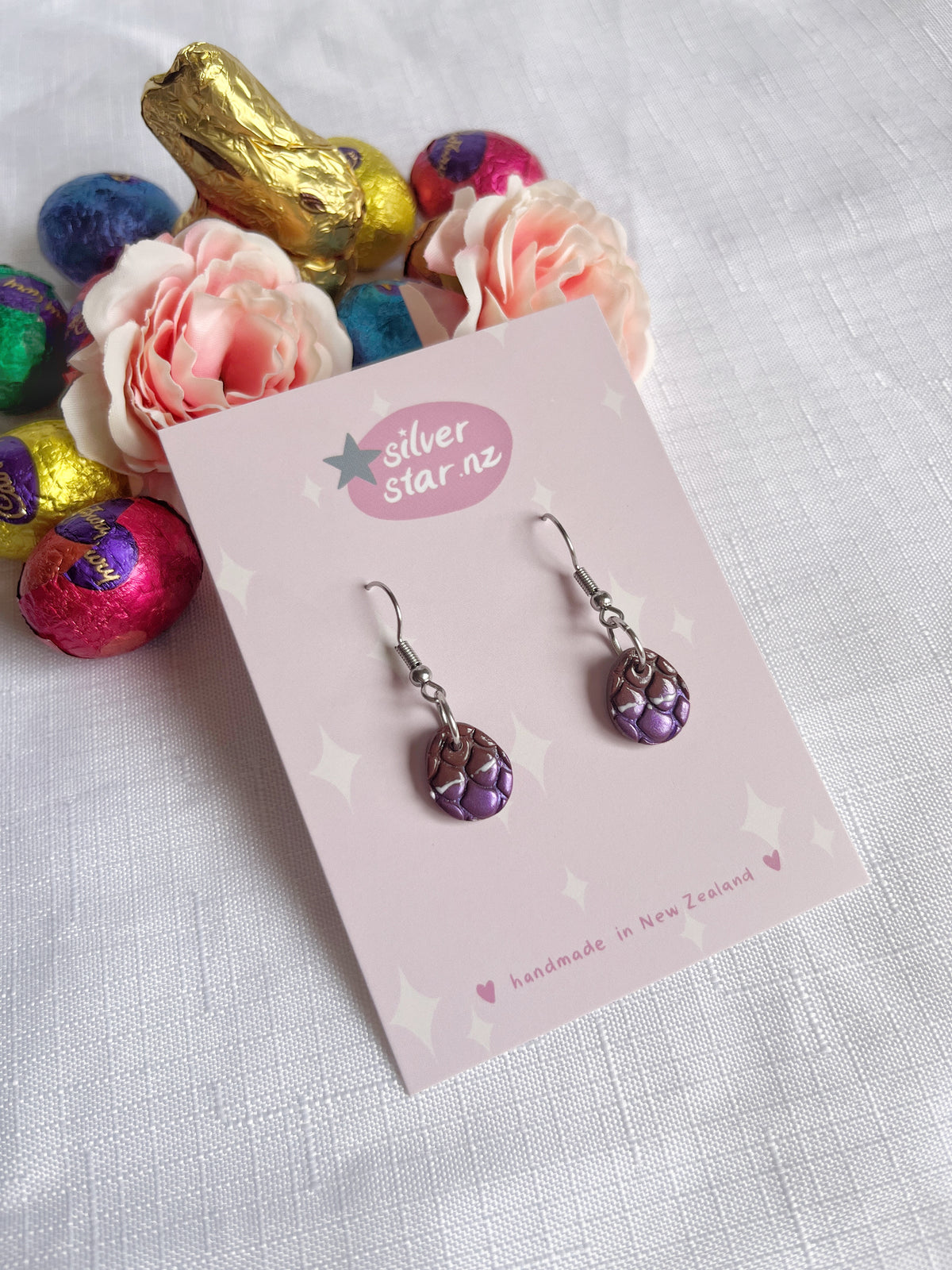The Opened Easter Egg Polymer Clay Earrings by silverstar.nz feature a unique handmade design with silver hooks and purple patterned beads. Displayed against the silverstar.nz card, these earrings amid colorful wrapped chocolates and pink flowers are crafted in New Zealand.