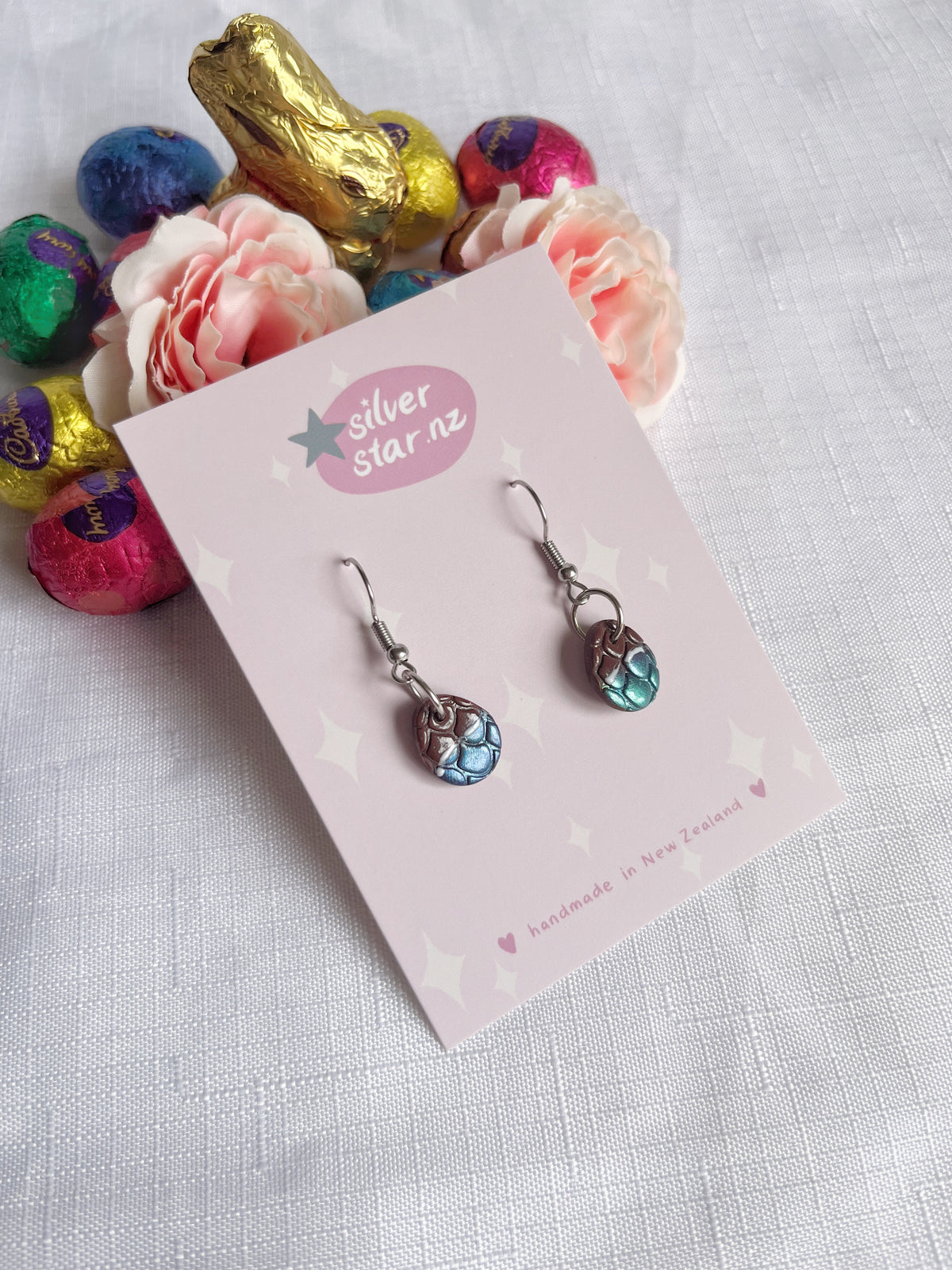 The Opened Easter Egg Polymer Clay Earrings by silverstar.nz showcase blue and silver textured beads on a card with pink flowers and colorful wrapped chocolate eggs on a textured white surface, adding unique artistry to any ensemble.