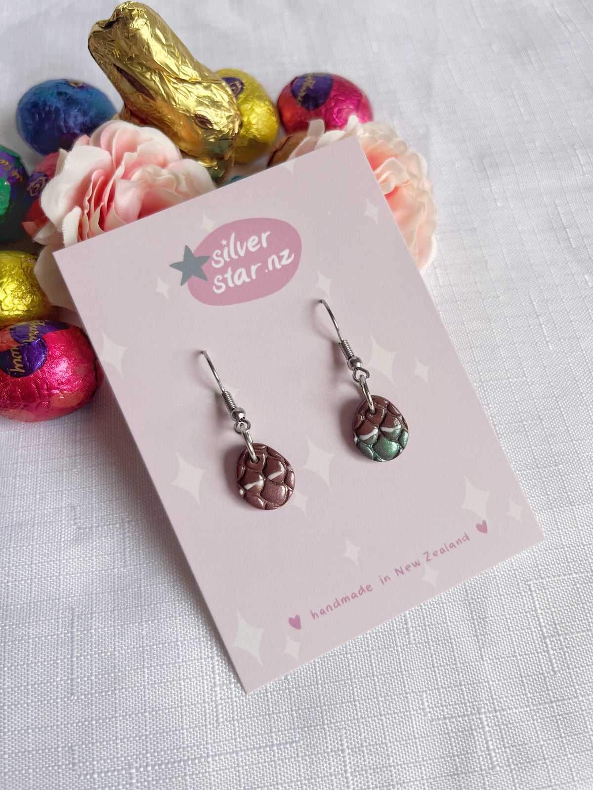 The Opened Easter Egg Polymer Clay Earrings by silverstar.nz feature a small, round, woven design. Displayed on a card labeled handmade in New Zealand, theyre set against pink roses and colorful foil-wrapped chocolate eggs on white fabric, adding a unique design touch.