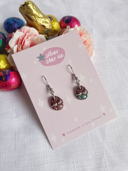 The Opened Easter Egg Polymer Clay Earrings by silverstar.nz feature a small, round, woven design. Displayed on a card labeled handmade in New Zealand, theyre set against pink roses and colorful foil-wrapped chocolate eggs on white fabric, adding a unique design touch.