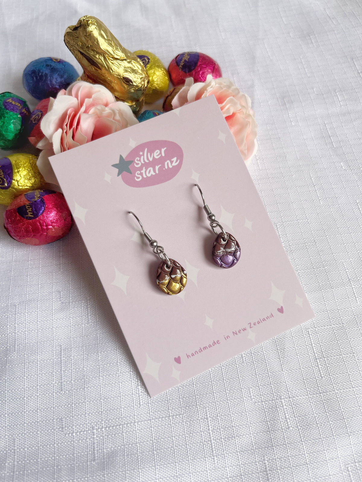 A pair of handmade dangle earrings, the Opened Easter Egg Polymer Clay Earrings, with metallic scales in gold and purple hues is displayed on a card labeled silverstar.nz. The background features a white cloth, pastel pink roses, and colorful foil-wrapped chocolates, including a gold bunny.