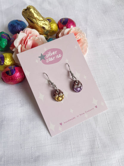 A pair of handmade dangle earrings, the Opened Easter Egg Polymer Clay Earrings, with metallic scales in gold and purple hues is displayed on a card labeled silverstar.nz. The background features a white cloth, pastel pink roses, and colorful foil-wrapped chocolates, including a gold bunny.