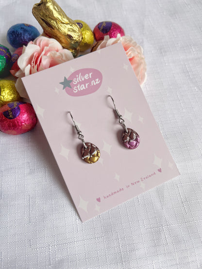 The handcrafted Opened Easter Egg earrings by silverstar.nz, showcasing purple and gold dragon-scale patterns, are displayed on a labeled card. The setup includes white cloth, pink roses, and vibrant foil-wrapped chocolates.