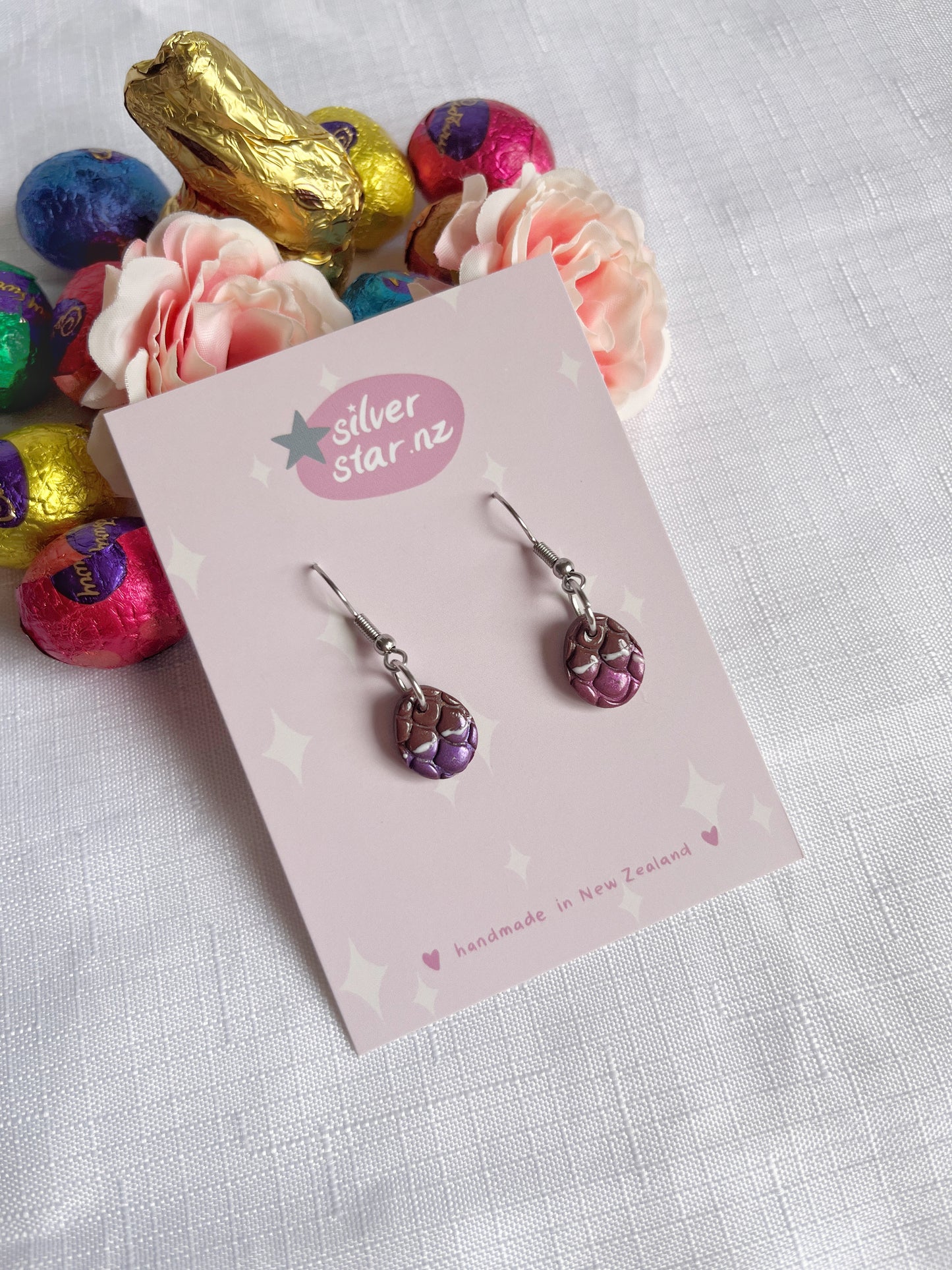 The Opened Easter Egg Polymer Clay Earrings by silverstar.nz feature a unique dragon-scale design displayed on their branded card. Set against a textured white surface, pink flowers and colorful wrapped candies enhance the presentation of these handmade earrings.