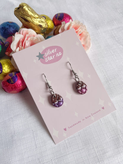 Displayed on a silverstar.nz card, the Opened Easter Egg Polymer Clay Earrings feature a handmade purple design with intricate craftsmanship, set against pink roses and colorful wrapped chocolates on white fabric by New Zealand artisans.