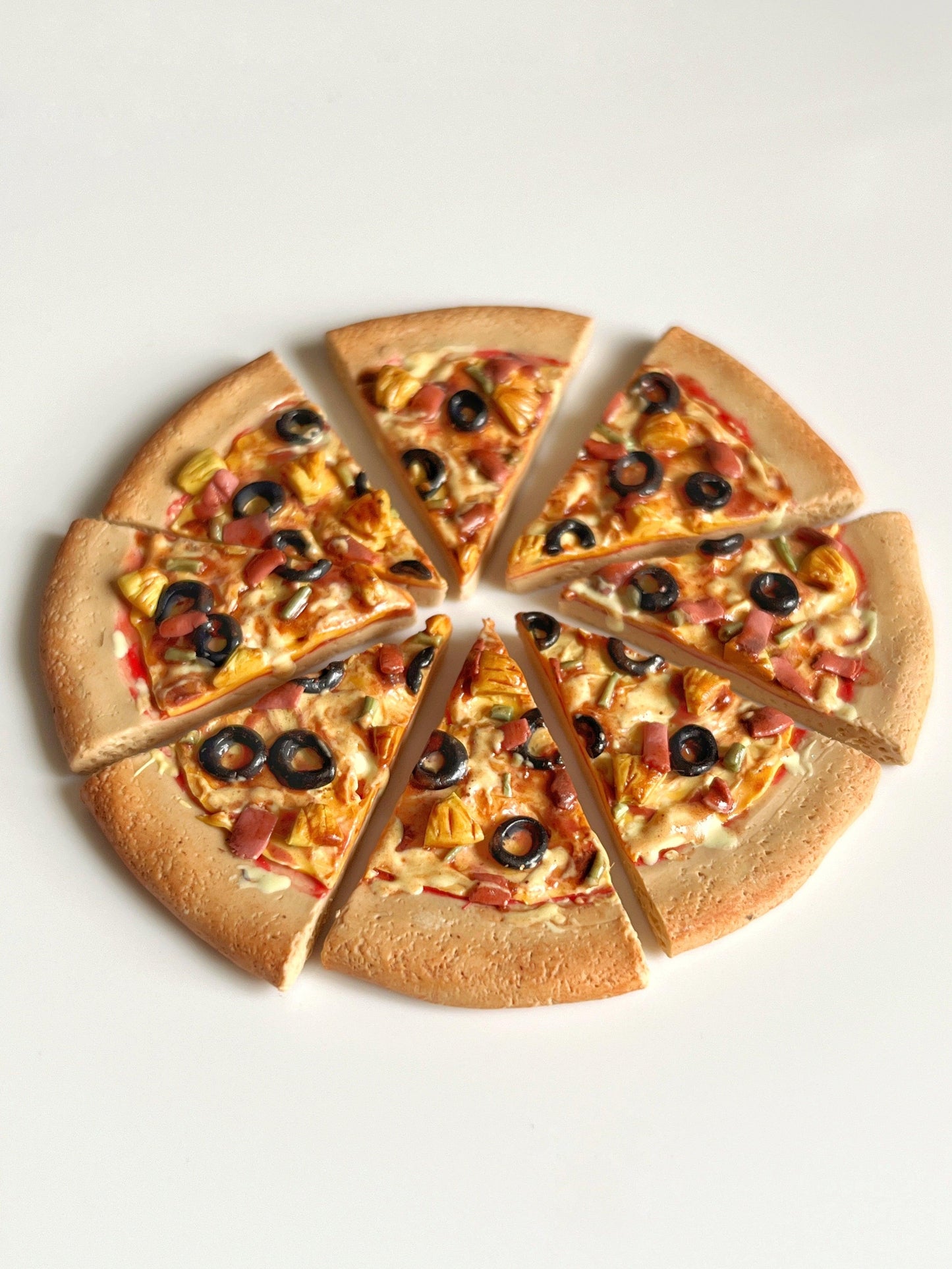 A pizza sliced into six pieces on a light background showcases black olives, pineapple chunks, corn, and bits of ham atop a golden brown crust. Much like the Pizza Polymer Clay Earrings, this pizza is both unique and delightful with intricate detail.