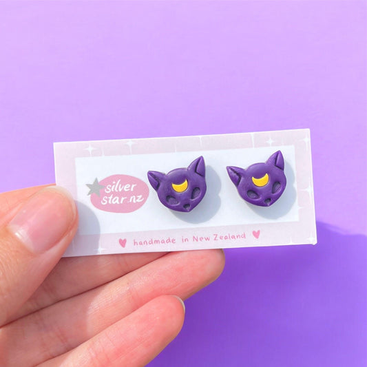 A hand holds a card reading "silverstar.nz" with heart illustrations, displaying Purple Moon Cat Polymer Clay Stud Earrings—purple cat-shaped studs with yellow crescent moons—set on a lavender background. Handmade in New Zealand.