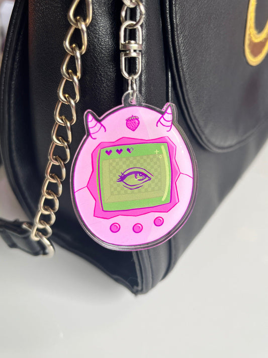 The Strawberry Tama Acrylic Keyring by silverstar.nz, featuring a pink virtual pet design with horns and an eye on-screen, hangs from a silver chain attached to a black bag.