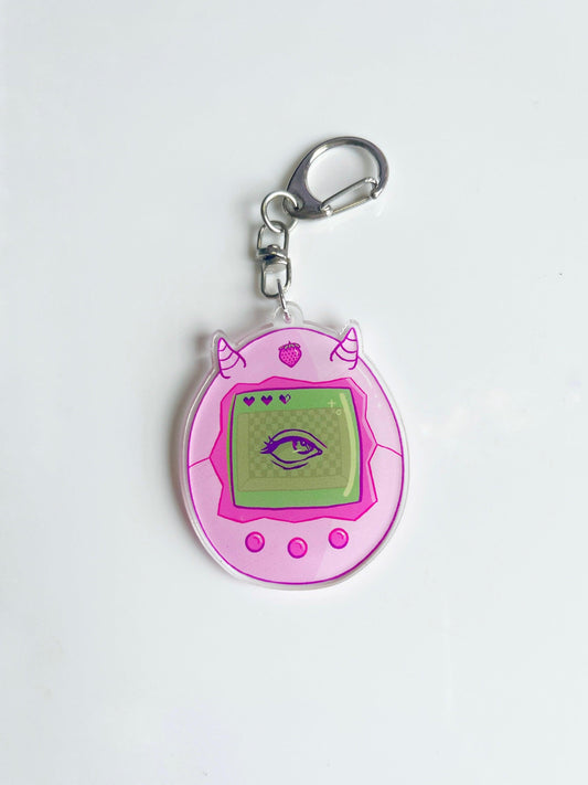 The Strawberry Tama Acrylic Keyring by silverstar.nz is a pink acrylic keyring shaped like a virtual pet with horns, featuring a green screen with an illustrated eye and heart icons. Includes a silver clip for easy attachment.