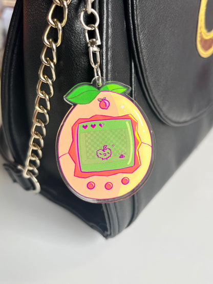 The Peach Tama Acrylic Keyring by silverstar.nz, artist-designed and peach-shaped, hangs from a black handbag’s chain strap. Its digital screen shows a pixelated pet with hearts above it, set against a white background.