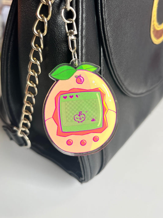 The Peach Tama Acrylic Keyring by silverstar.nz, artist-designed and peach-shaped, hangs from a black handbag’s chain strap. Its digital screen shows a pixelated pet with hearts above it, set against a white background.