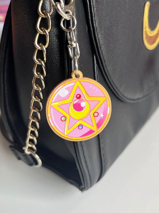 A close-up of a black purse with a silver chain strap, featuring the Moon Locket Acrylic Keyring by silverstar.nz—a vibrant pink and gold charm shaped like a magical star and crescent moon.