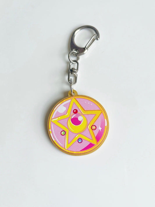 The Moon Locket Acrylic Keyring by silverstar.nz is a round acrylic keychain with a silver clasp, featuring a pink and yellow star design with colorful gems inspired by magical girl anime—a perfect accessory for any fan.
