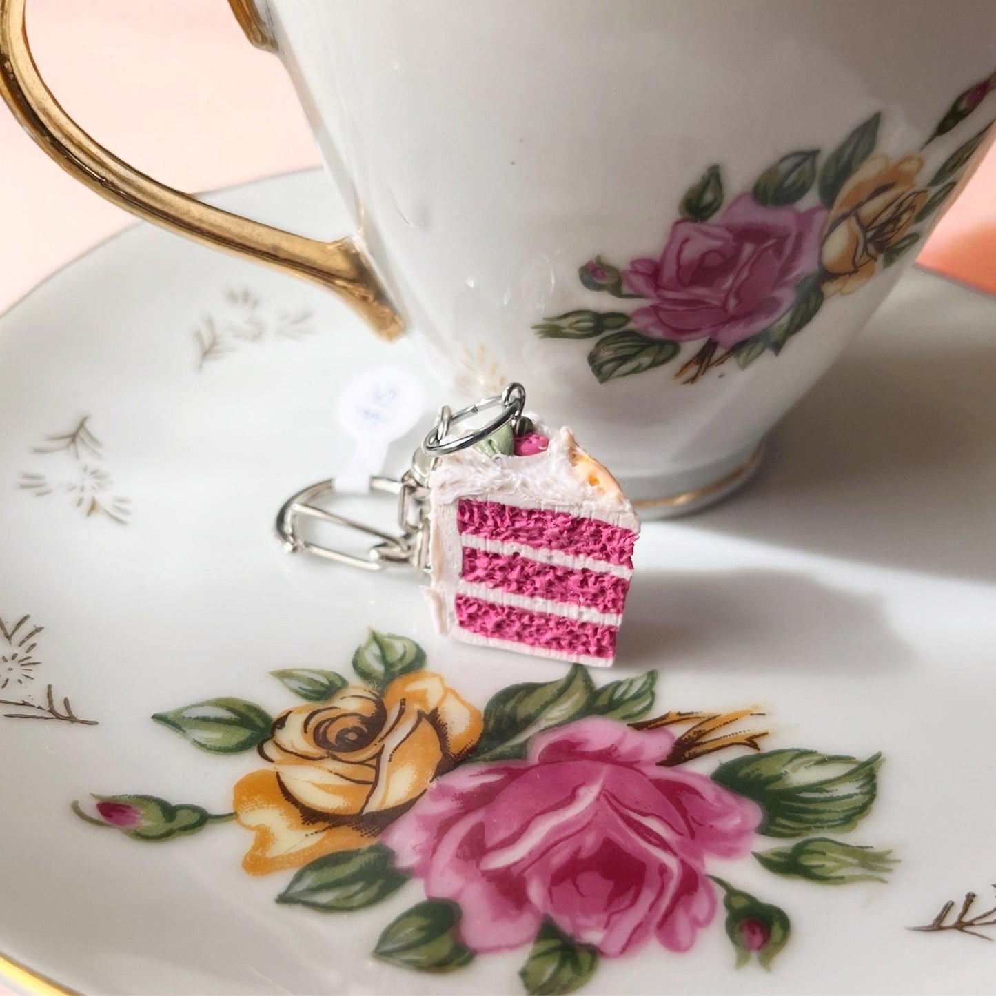 A Strawberry Cake Polymer Clay Keyring, featuring a pink layered cake charm made from durable polymer clay, rests on a white porcelain plate adorned with floral designs. In the background, a matching teacup with gold detailing exhibits a distinct funky design.