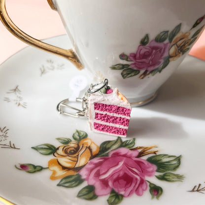A Strawberry Cake Polymer Clay Keyring, featuring a pink layered cake charm made from durable polymer clay, rests on a white porcelain plate adorned with floral designs. In the background, a matching teacup with gold detailing exhibits a distinct funky design.