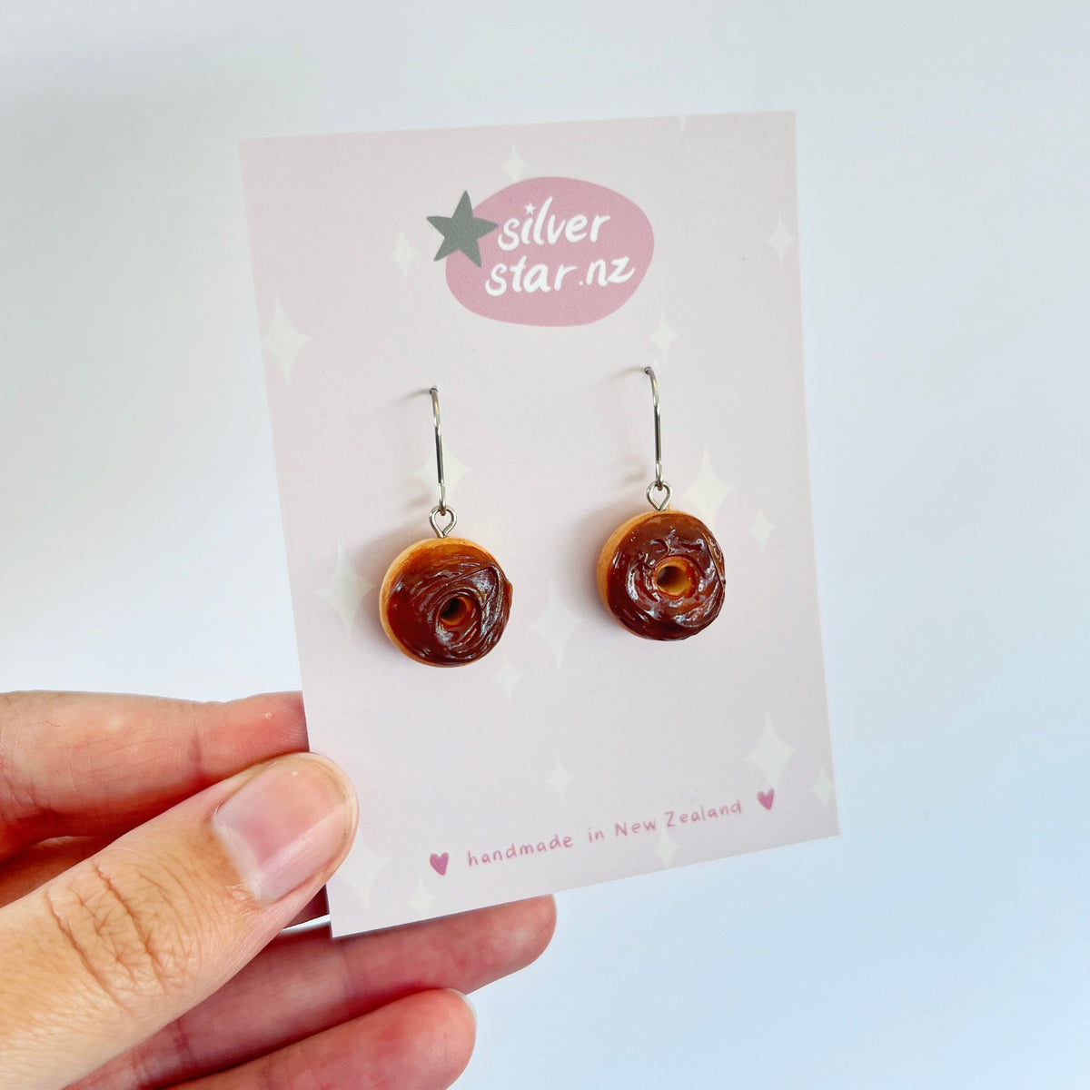 Close-up of a hand holding a card with unique polymer clay earrings shaped like chocolate iced donuts. The card features a silverstar.nz logo and the text "handmade in New Zealand." These handmade earrings have hooks for pierced ears.