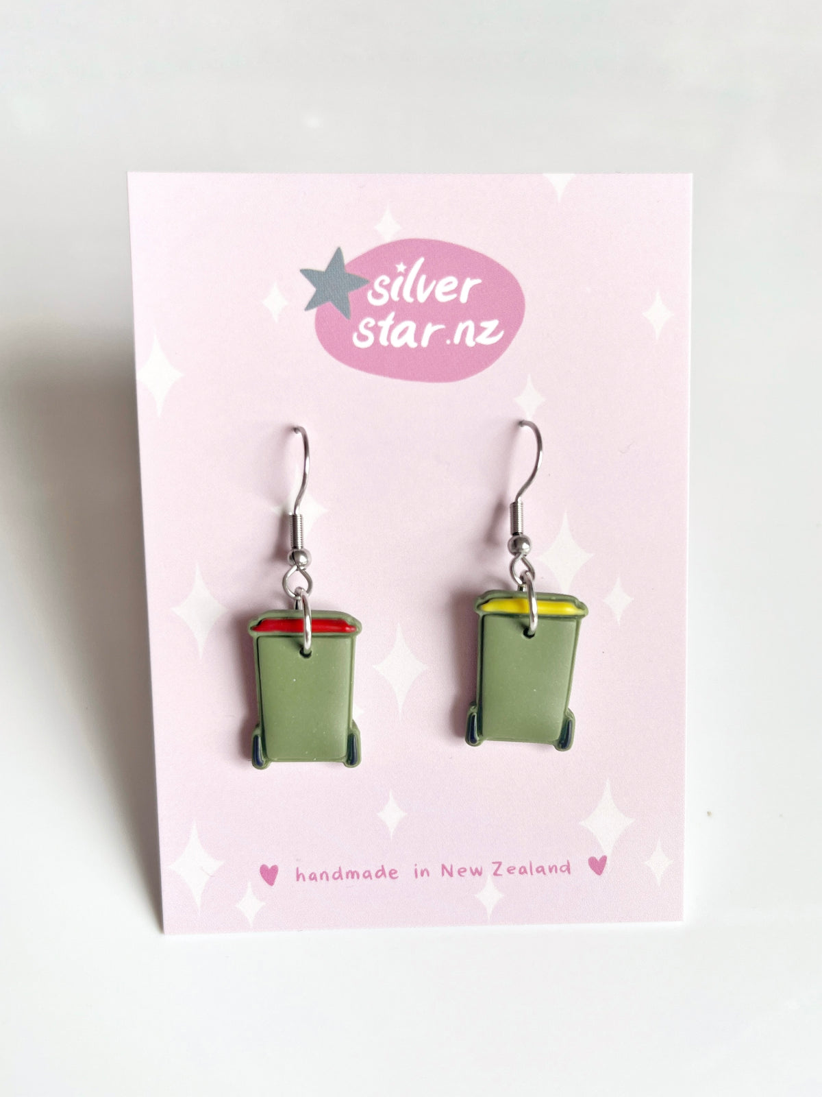 The Rubbish Bin Polymer Clay Earrings by silverstar.nz are shaped like green wheelie bins with red and yellow lids, presented on a pink card with stars. Handmade in New Zealand, these unique earrings capture attention.