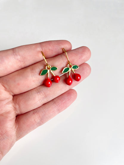 A hand displays the Mini Cherry Enamel Earrings from silverstar.nz, featuring vibrant red cherries with green leaves. These charming earrings have gold hooks and are set against a light gray background. Crafted with handmade polymer clay for a unique touch.